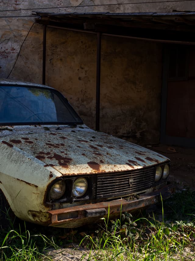 An old, rusted car is parked in front of a weathered building, surrounded by overgrown grass